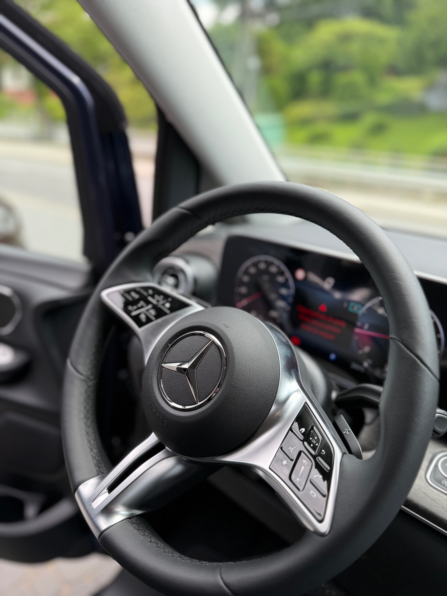 Wnętrze samochodu Mercedes: kierownica z logo, deska rozdzielcza z wyświetlaczem i widok na zieleń za oknem. Nowoczesny design i ergonomia.