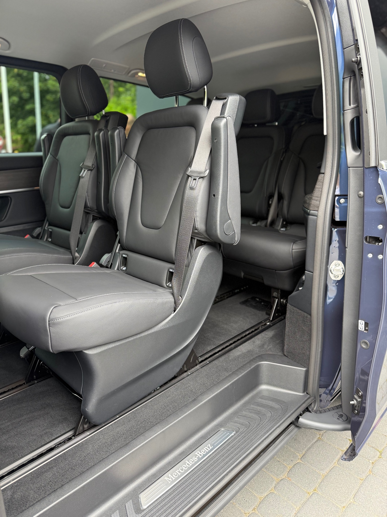 Wnętrze komfortowego busa Mercedes-Benz z czarnymi skórzanymi fotelami, widok przez otwarte drzwi boczne. Widoczny system mocowania foteli.