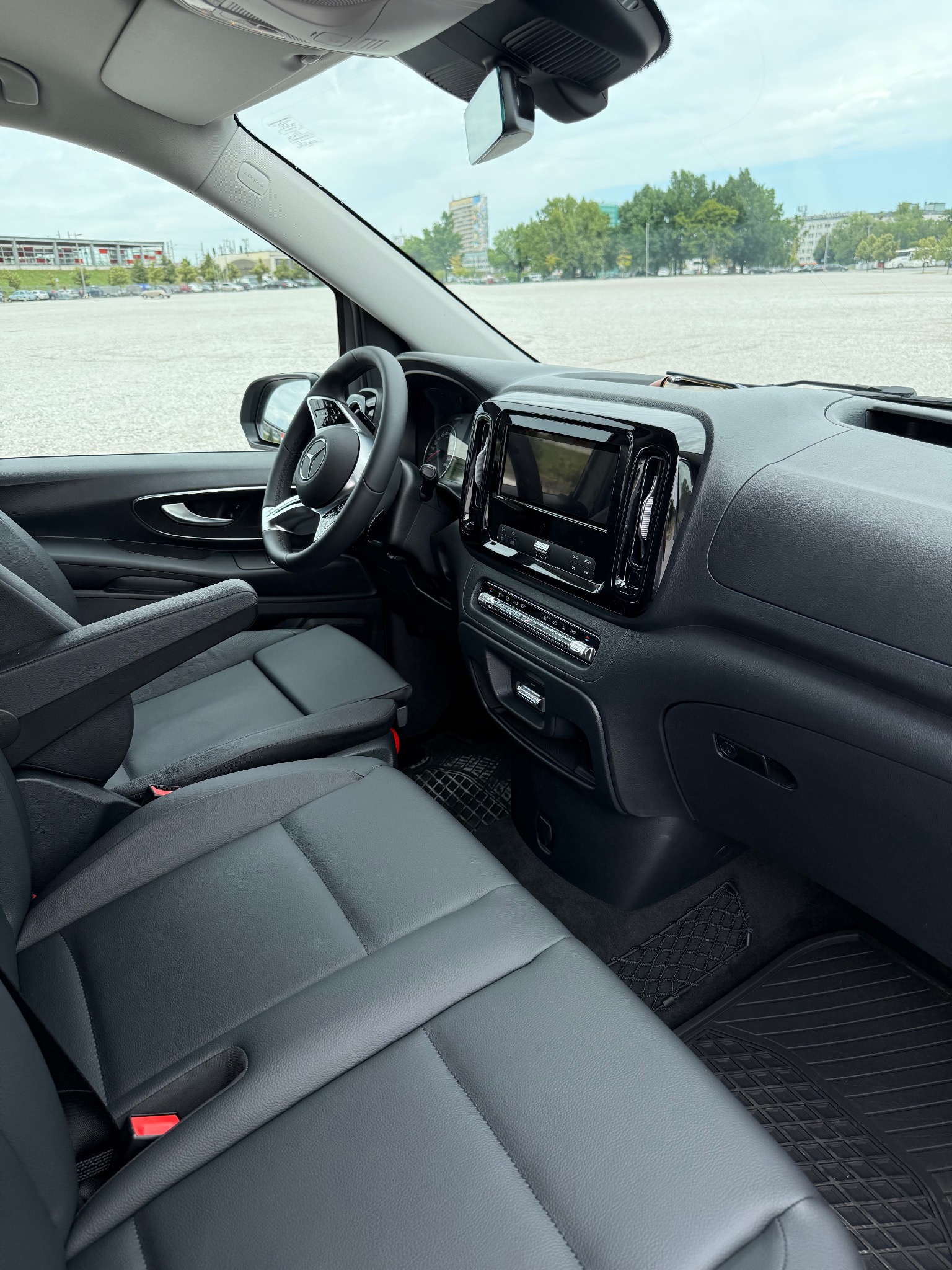 Wnętrze busa Mercedes Vito: czarna skórzana tapicerka, deska rozdzielcza z ekranem dotykowym, widok na parking i budynki w tle przez szybę. Detal.