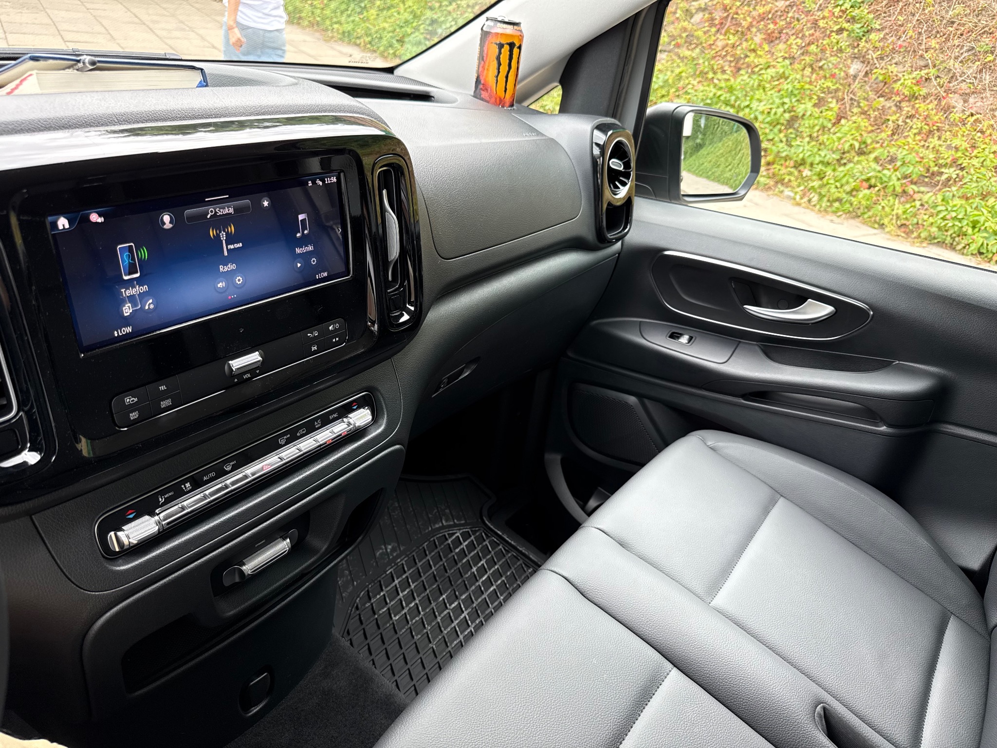 Wnętrze samochodu z szarymi skórzanymi siedzeniami, widoczny ekran dotykowy i puszka napoju na desce rozdzielczej. Widok z perspektywy pasażera.