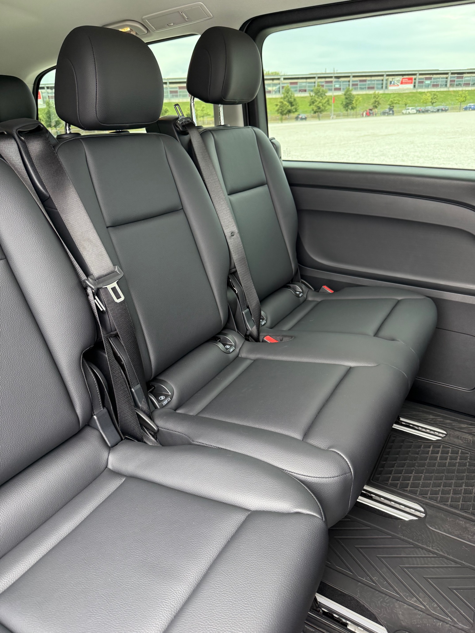 Czarne, skórzane fotele z pasami bezpieczeństwa w przestronnym wnętrzu busa, widok z boku, za oknem parking z zielenią. Detal wnętrza pojazdu.