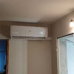 Biały klimatyzator marki AUX zamontowany na ścianie w pokoju z beżową tapetą, widoczny wyświetlacz z temperaturą 24 stopnie Celsjusza, obok drzwi z surowym betonowym wykończeniem ościeżnicy...