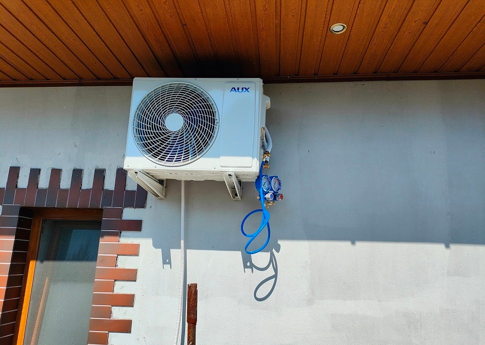 Biały klimatyzator marki AUX zamontowany na szarej ścianie zewnętrznej domu, z podłączonymi niebieskimi wężami manometru serwisowego.