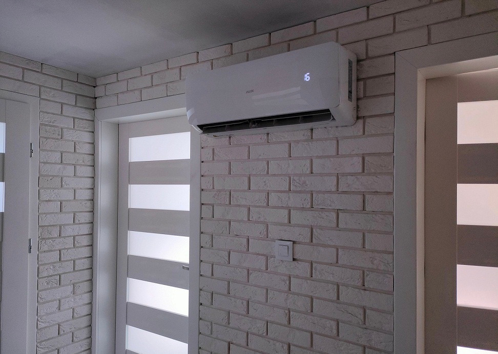 Biały klimatyzator ścienny z wyświetlaczem temperatury na tle ściany z białej cegły, widoczny włącznik światła i fragmenty białych drzwi.