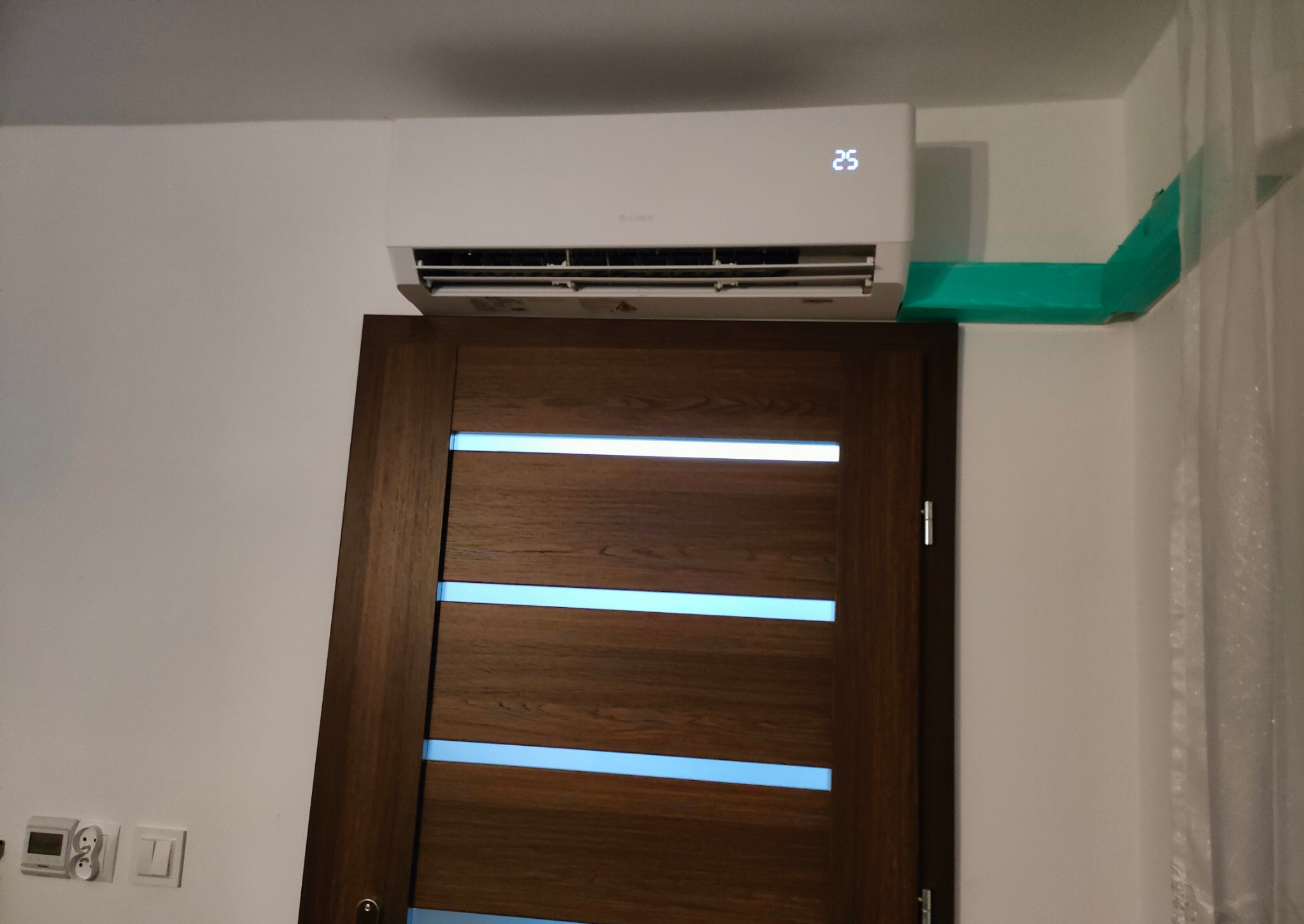 Biały klimatyzator zamontowany nad drewnianymi drzwiami z podświetleniem LED, z wyświetlaczem temperatury ustawionym na 25 stopni Celsjusza, rura odprowadzająca zabezpieczona zieloną folią ochronną.