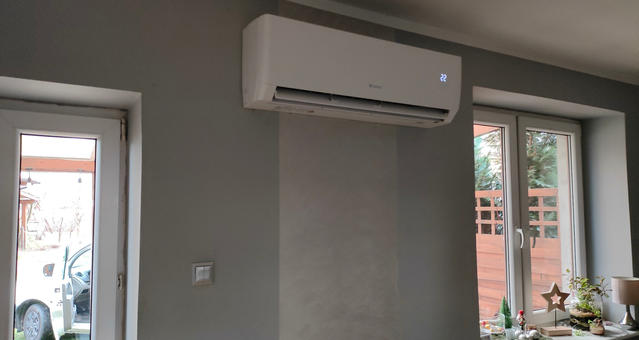 Biały klimatyzator wiszący na szarej ścianie w pokoju z oknami, na wyświetlaczu klimatyzatora widoczna temperatura 22 stopnie.