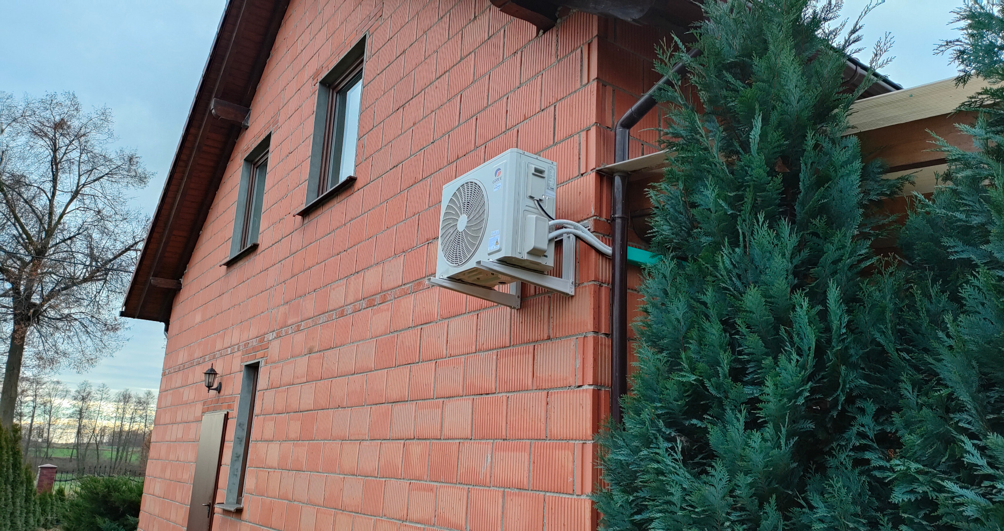 Zewnętrzna jednostka klimatyzacji marki Gree zamontowana na ceglanej ścianie domu, z widocznymi przewodami i wspornikami mocującymi, częściowo zasłonięta przez iglaste drzewo.