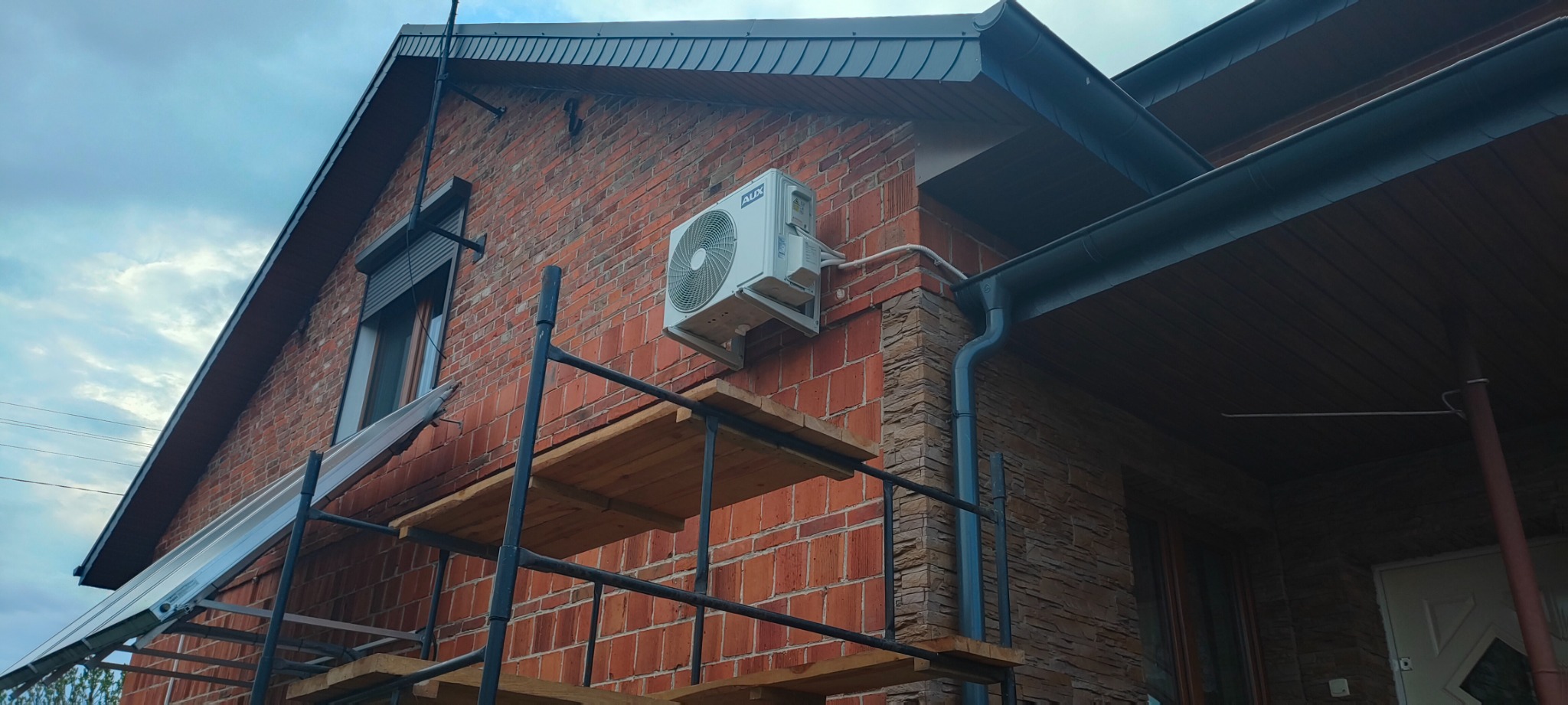 Biały klimatyzator marki AUX zamontowany na ceglanej ścianie domu, widoczny z rusztowania, z rurami odprowadzającymi skropliny.