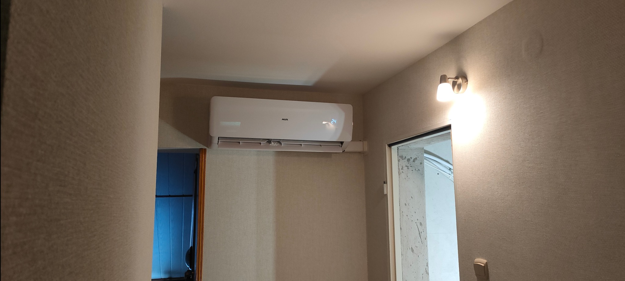 Biały klimatyzator marki AUX zamontowany na ścianie w pokoju z beżową tapetą, widoczny wyświetlacz z temperaturą 24 stopnie Celsjusza, obok drzwi z surowym betonowym wykończeniem ościeżnicy...