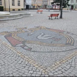 Herb miasta ułożony z kostki granitowej na placu miejskim, z fontanną i ławkami w tle.