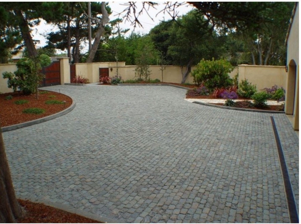 Szeroki podjazd wyłożony szarą kostką granitową, z obrzeżami i zielenią w tle oraz drewnianą bramą w oddali.