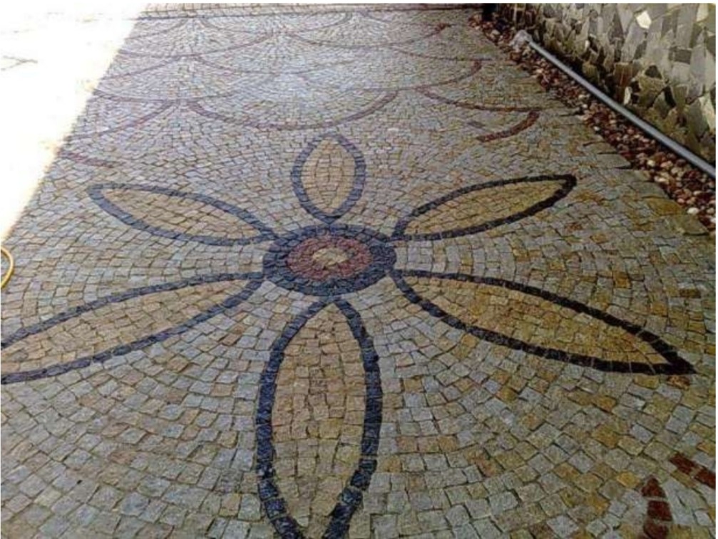 Ułożona kostka granitowa w kształcie kwiatu na podjeździe, z widocznym fragmentem ściany i rynny w tle.