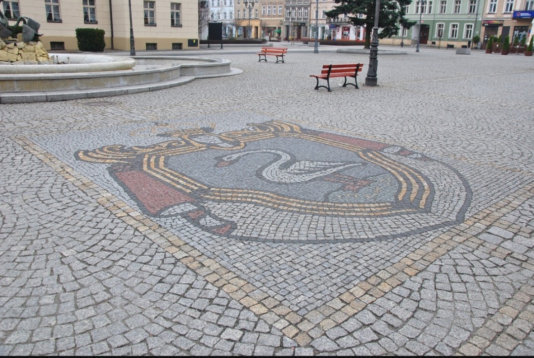 Herb miasta ułożony z kostki granitowej na placu miejskim, z fontanną i ławkami w tle.