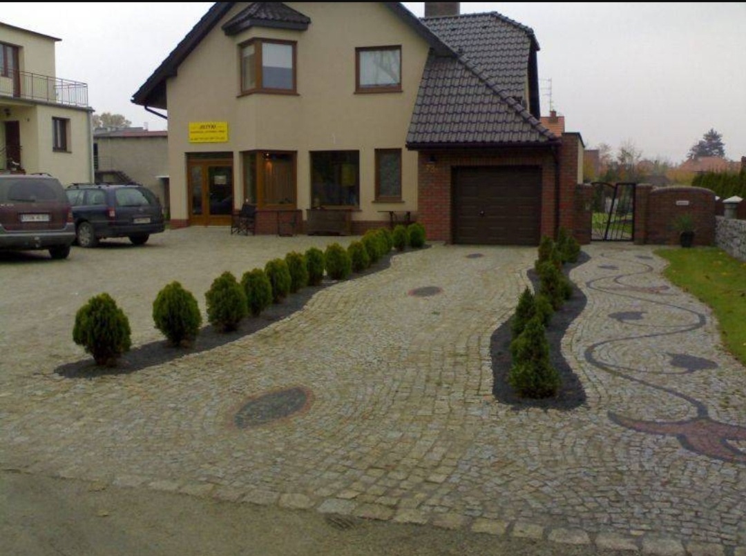 Podjazd do domu wyłożony kostką granitową w odcieniach szarości, z dekoracyjnymi wstawkami z ciemniejszego kamienia tworzącymi wzory i obwódki z nasadzeniami iglaków po bokach.