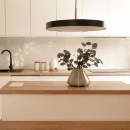 Nowoczesna kuchnia z wyspą, białymi szafkami i heksagonalnymi płytkami na ścianie. Na wyspie wazon z zielonymi gałązkami i czarna lampa sufitowa.