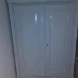 Odnowiona, biała szafa dwudrzwiowa z dwiema szufladami, widoczne ślady świeżego malowania z połyskiem, ciemne uchwyty szuflad.