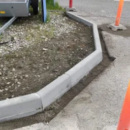 Świeżo ułożony betonowy obrzeże chodnika z widocznymi śladami ziemi i piasku, pomarańczowe pachołki ostrzegawcze w tle, przyczepka samochodowa po lewej stronie.