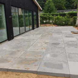 Nowo ułożona, duża, szara kostka brukowa tworząca taras przy nowoczesnym domu z czarną elewacją i dużymi oknami przesuwnymi, obrzeża tarasu obsypane piaskiem.