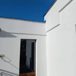 Biała elewacja budynku z drewnianą ramą okienną, odbijająca wnętrze w szybie, na tle błękitnego nieba.