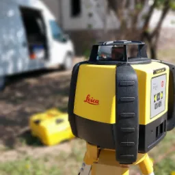 Żółty laser rotacyjny Leica Rugby 640 na statywie, z białym vanem w tle na trawie.