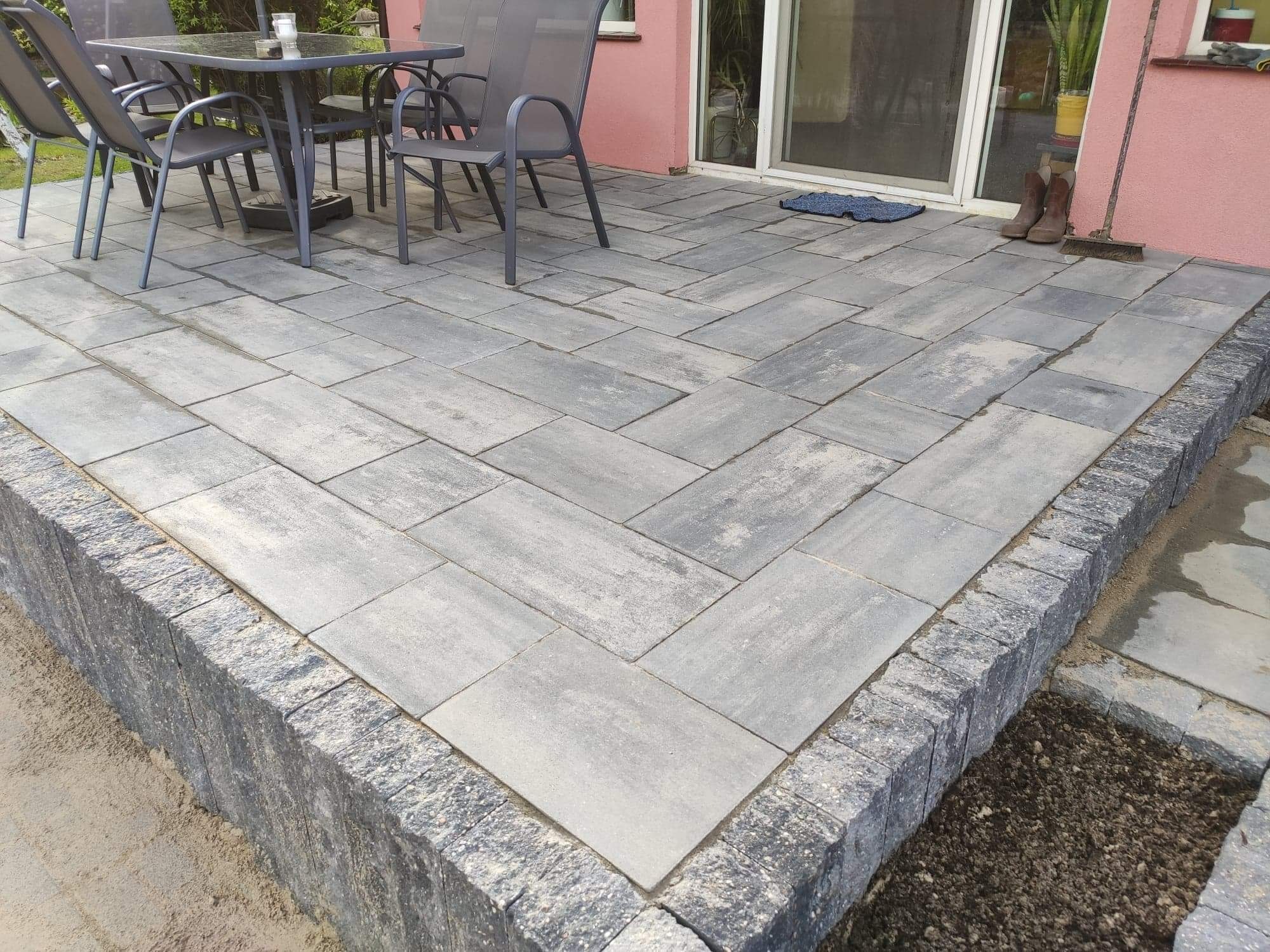 Szara kostka brukowa ułożona w jodełkę na tarasie przydomowym, z szarymi meblami ogrodowymi i butami roboczymi przy ścianie budynku.