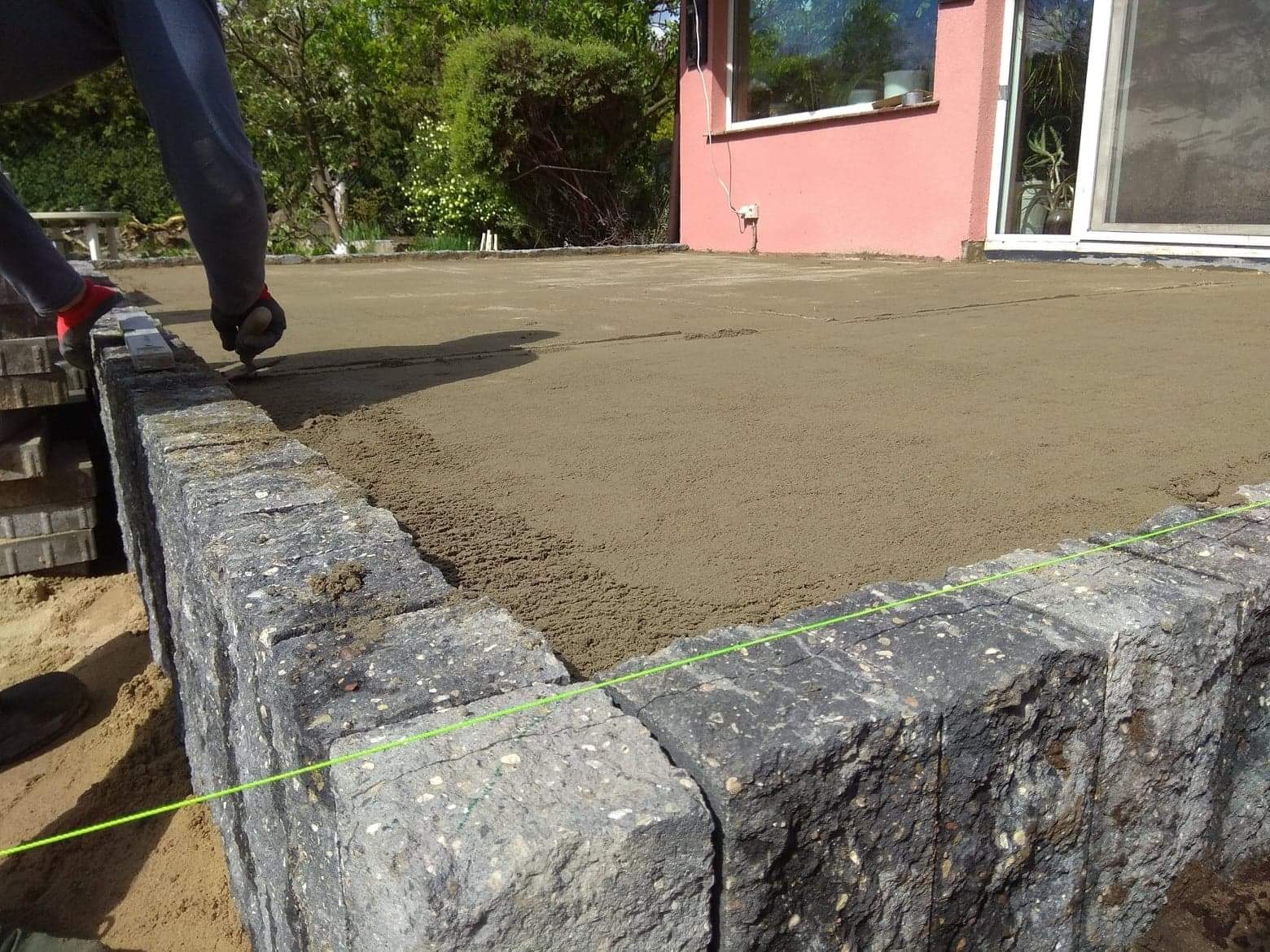 Przygotowanie podłoża pod kostkę brukową: wyrównywanie piasku na tarasie przylegającym do domu z różową elewacją, widoczny fragment murku oporowego z szarego kamienia.