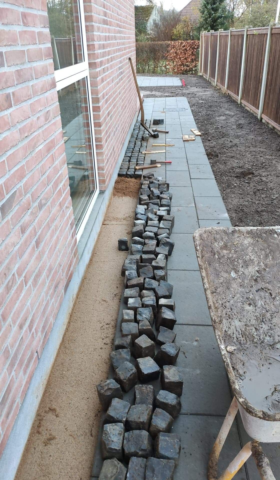 Przygotowanie podłoża i układanie kostki granitowej wzdłuż ściany budynku, widoczny piasek, narzędzia brukarskie i taczka z zaprawą.