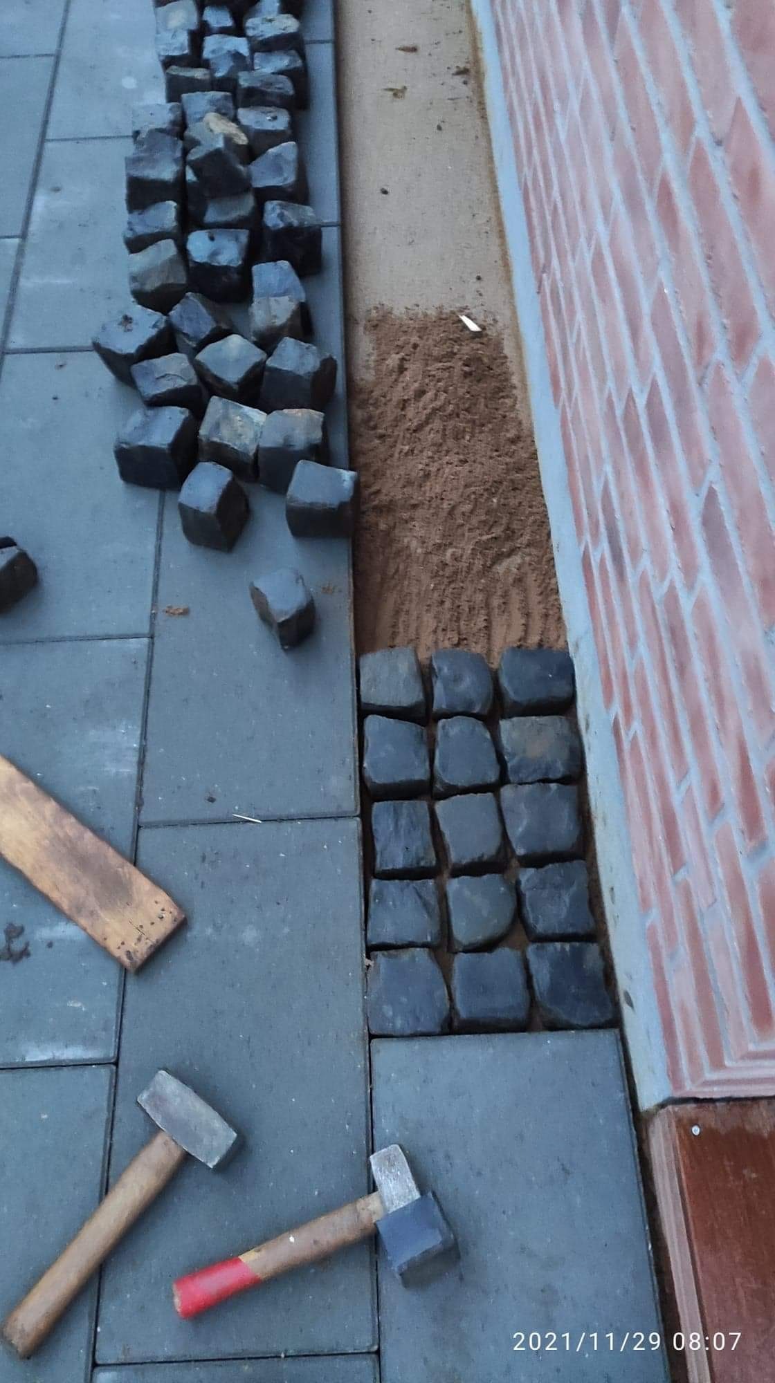 Fragment układania kostki granitowej przy ścianie budynku, widoczne dwa młotki brukarskie i drewniana listwa