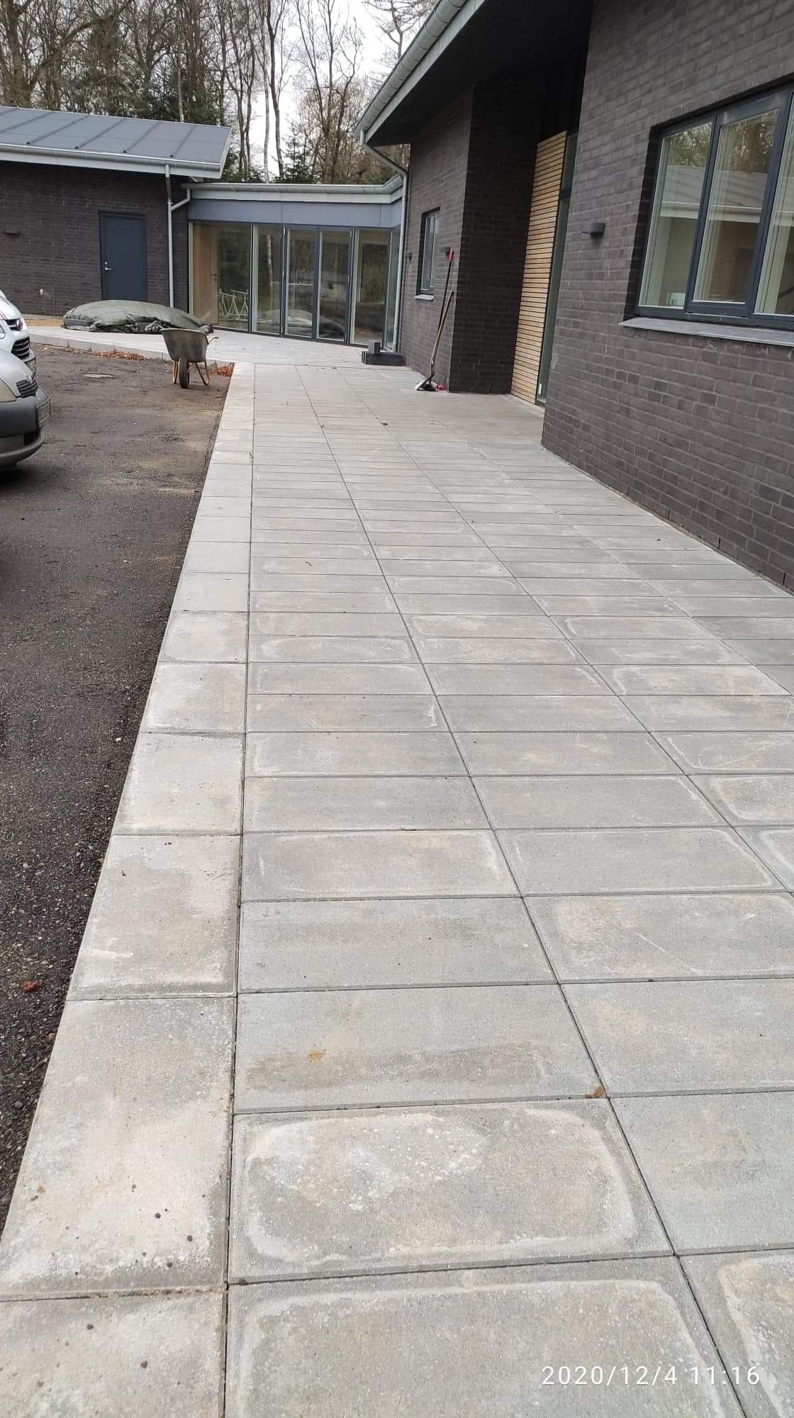 Ułożona, prostokątna, jasnoszara kostka brukowa tworząca chodnik przed nowoczesnym domem z ciemnej cegły; widoczny fragment samochodu, taczka i budynek gospodarczy w tle.