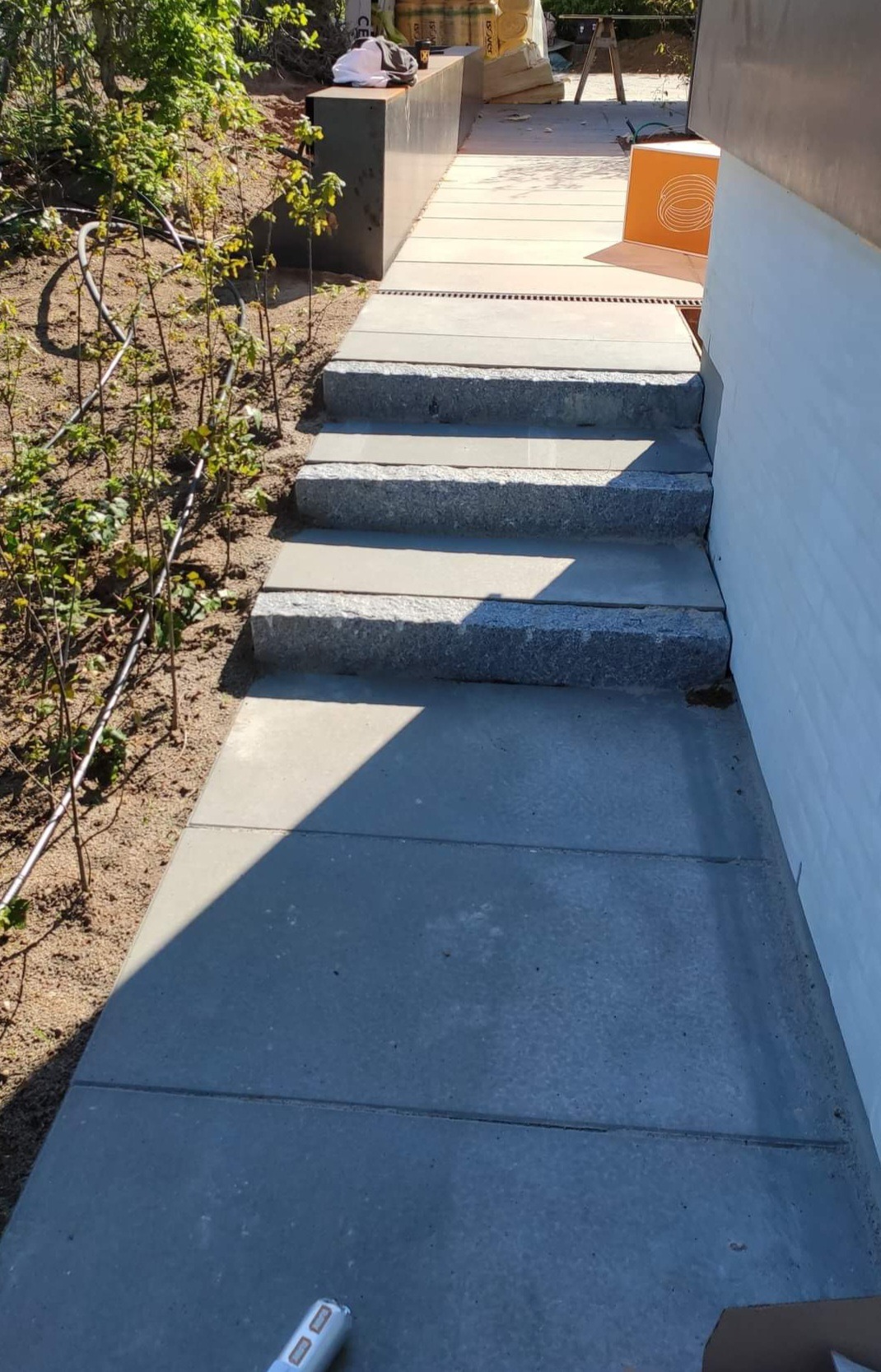 Nowoczesne podejście do domu z szarej kostki brukowej i kamiennych schodów, z widocznym systemem odwadniającym i elementami ogrodu w tle.