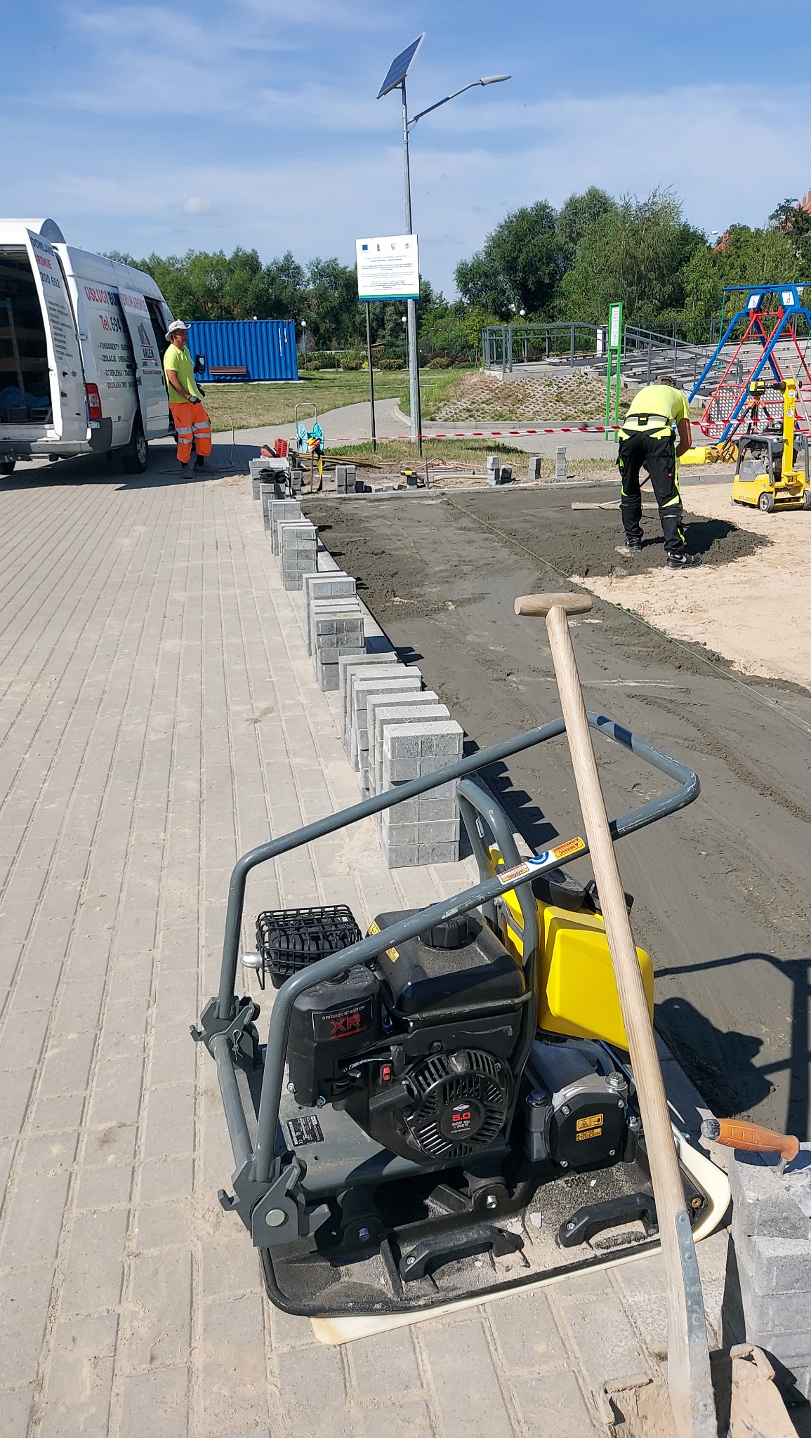 Układanie kostki brukowej na placu zabaw: widoczna ubijarka, pracownicy w odblaskowych ubraniach, częściowo ułożona kostka i plac zabaw w tle.