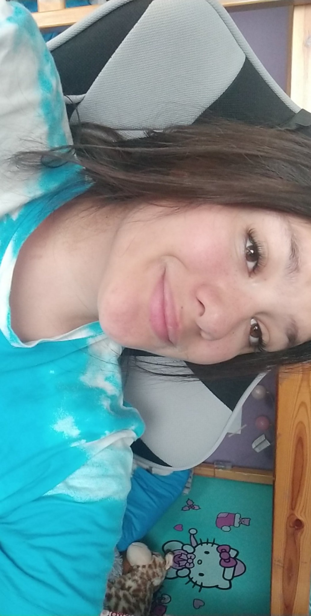 Uśmiechnięta dziewczyna leżąca na łóżku z turkusową pościelą tie-dye i pluszowym żyrafą, na tle dywanik z Hello Kitty.