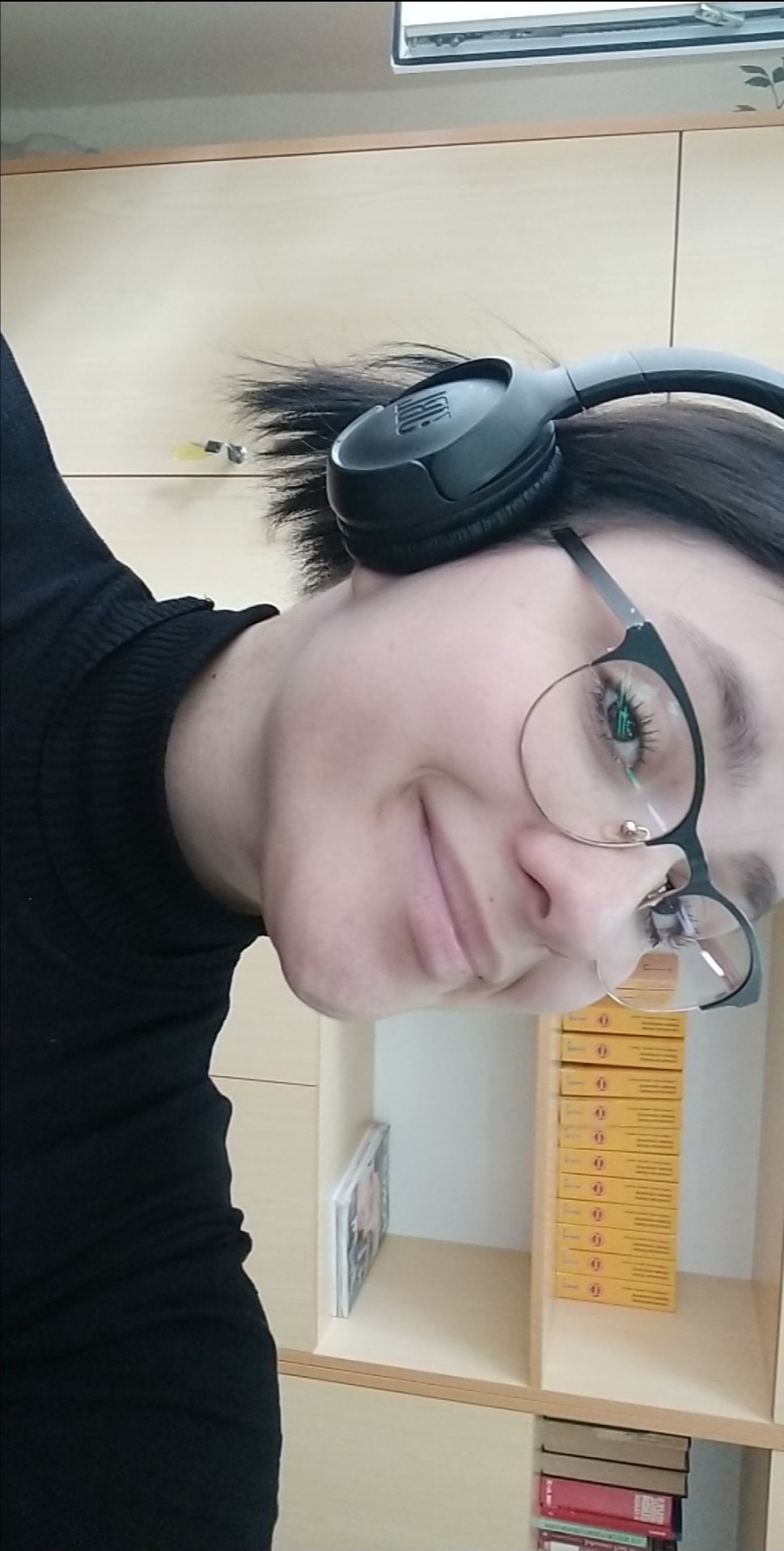 Portret kobiety w okularach i słuchawkach JBL, uśmiechającej się do kamery, na tle półki z książkami i pudełkami.