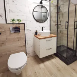 Nowoczesna łazienka z kabiną prysznicową typu walk-in z czarnymi profilami, okrągłym lustrem i umywalką nablatową na szafce z drewnianym blatem.