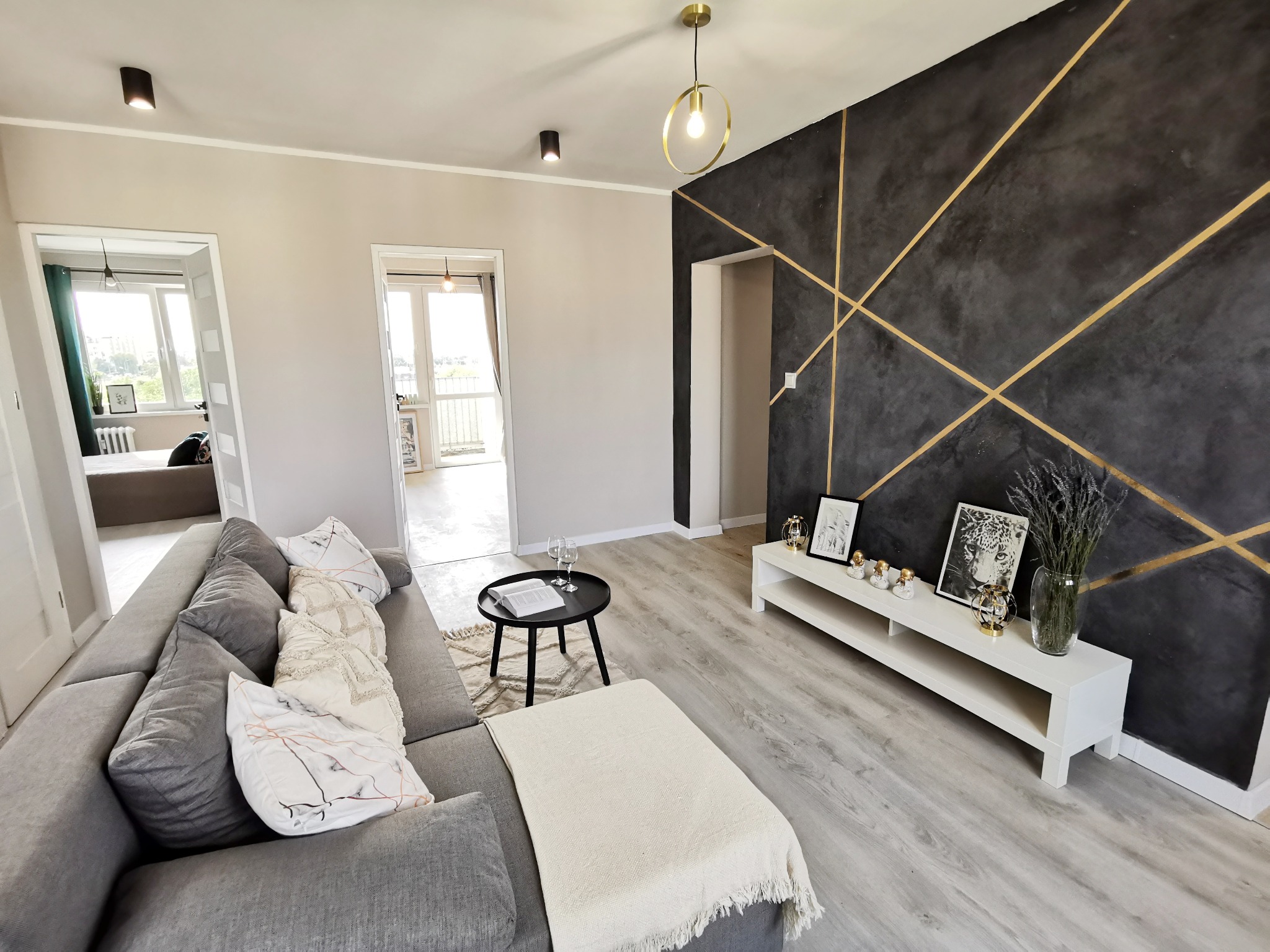Jasne wnętrze salonu z szarą sofą, czarnym stolikiem kawowym i ścianą z dekoracyjnym, ciemnym tynkiem z geometrycznym wzorem w złotym kolorze.