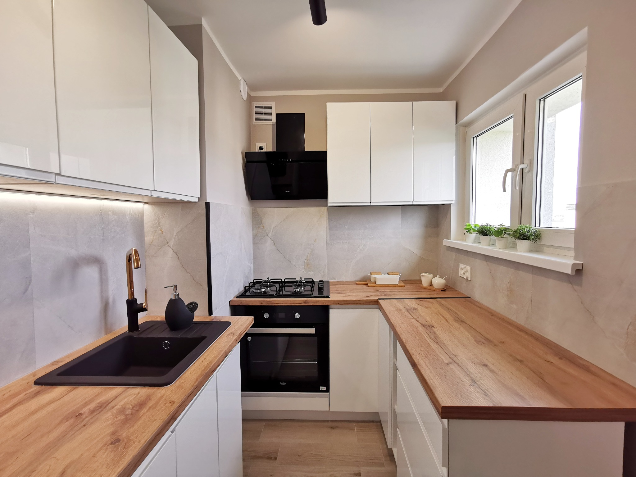 Jasna kuchnia z białymi szafkami, drewnianym blatem i czarną armaturą, widok na okno z roślinami.