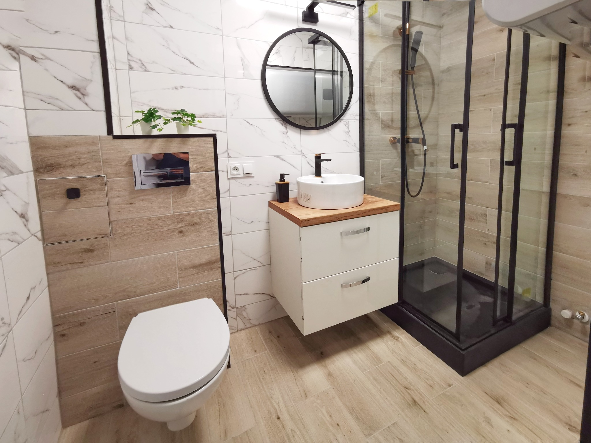 Nowoczesna łazienka z kabiną prysznicową typu walk-in z czarnymi profilami, okrągłym lustrem i umywalką nablatową na szafce z drewnianym blatem.