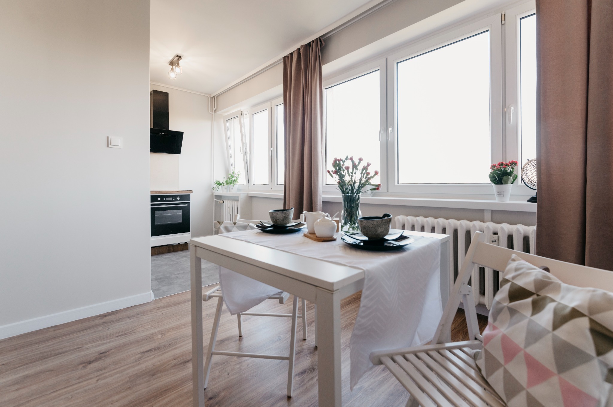 Jasne wnętrze mieszkania z drewnianą podłogą, białym stołem nakrytym do posiłku, białymi oknami z brązowymi zasłonami oraz widoczną kuchnią w tle.