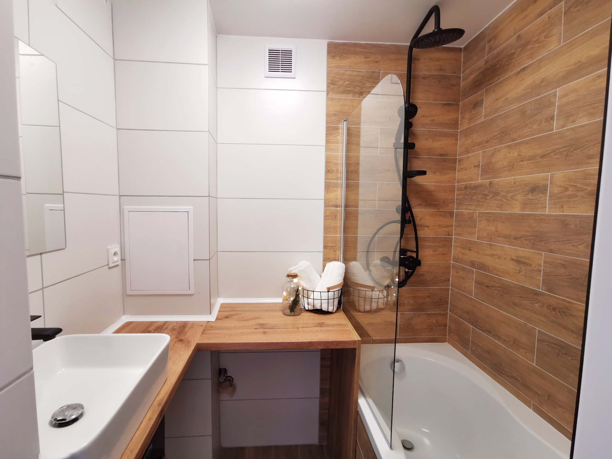 Nowoczesna łazienka z białymi i drewnopodobnymi płytkami, czarną armaturą prysznicową, umywalką nablatową i drewnianym blatem.