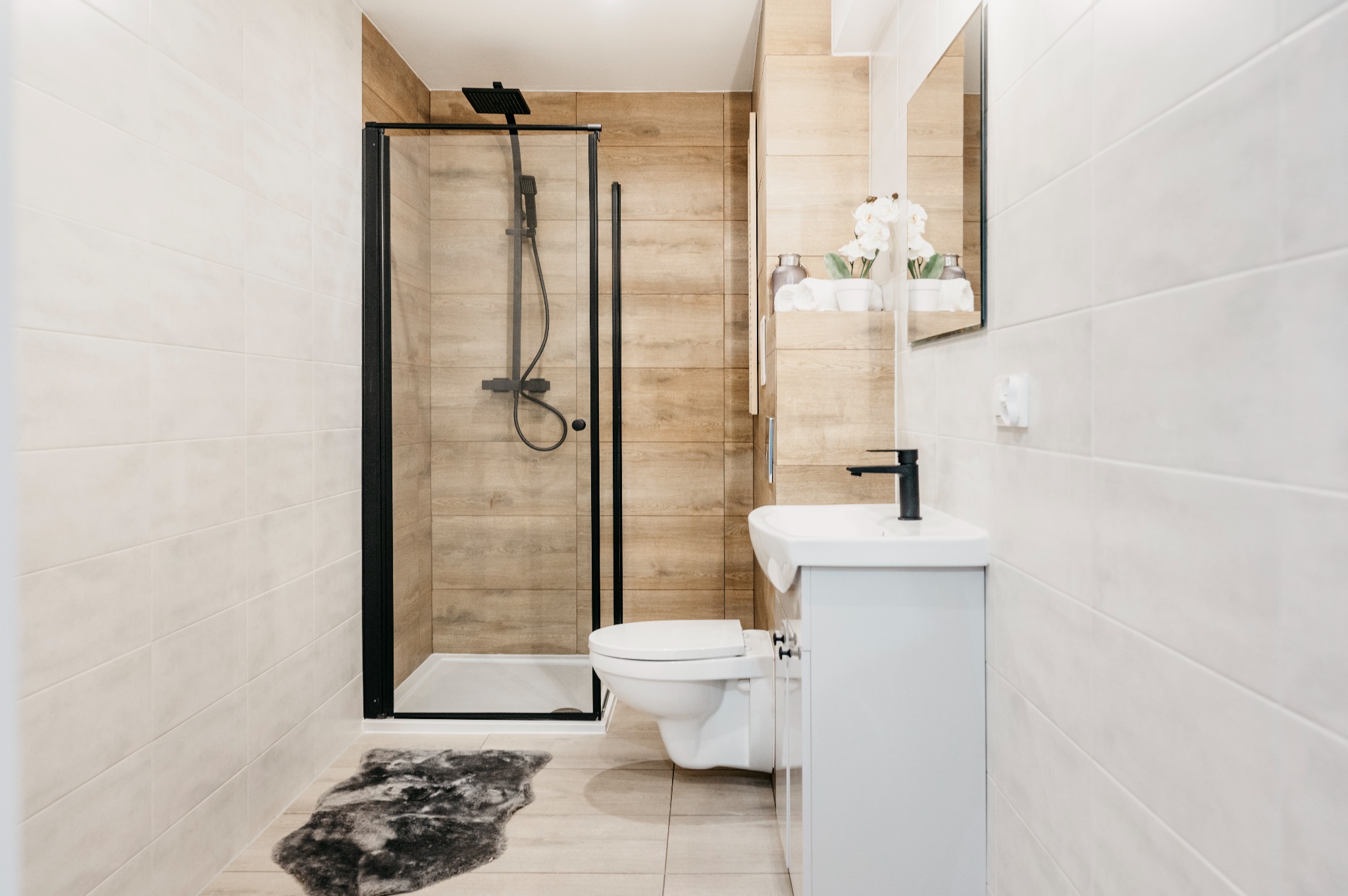 Nowoczesna łazienka z kabiną prysznicową typu walk-in z czarnymi ramami, umywalką nablatową z szafką w kolorze białym i toaletą podwieszaną, wykończona płytkami imitującymi drewno i jasnymi...