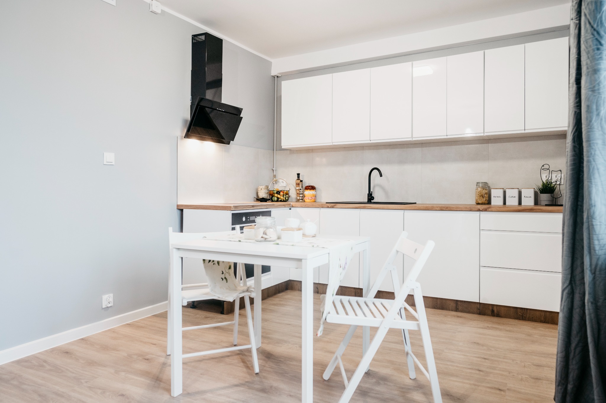 Jasna kuchnia z białymi szafkami, drewnianym blatem i czarnym okapem, uzupełniona białym stołem z krzesłami i panelami podłogowymi.