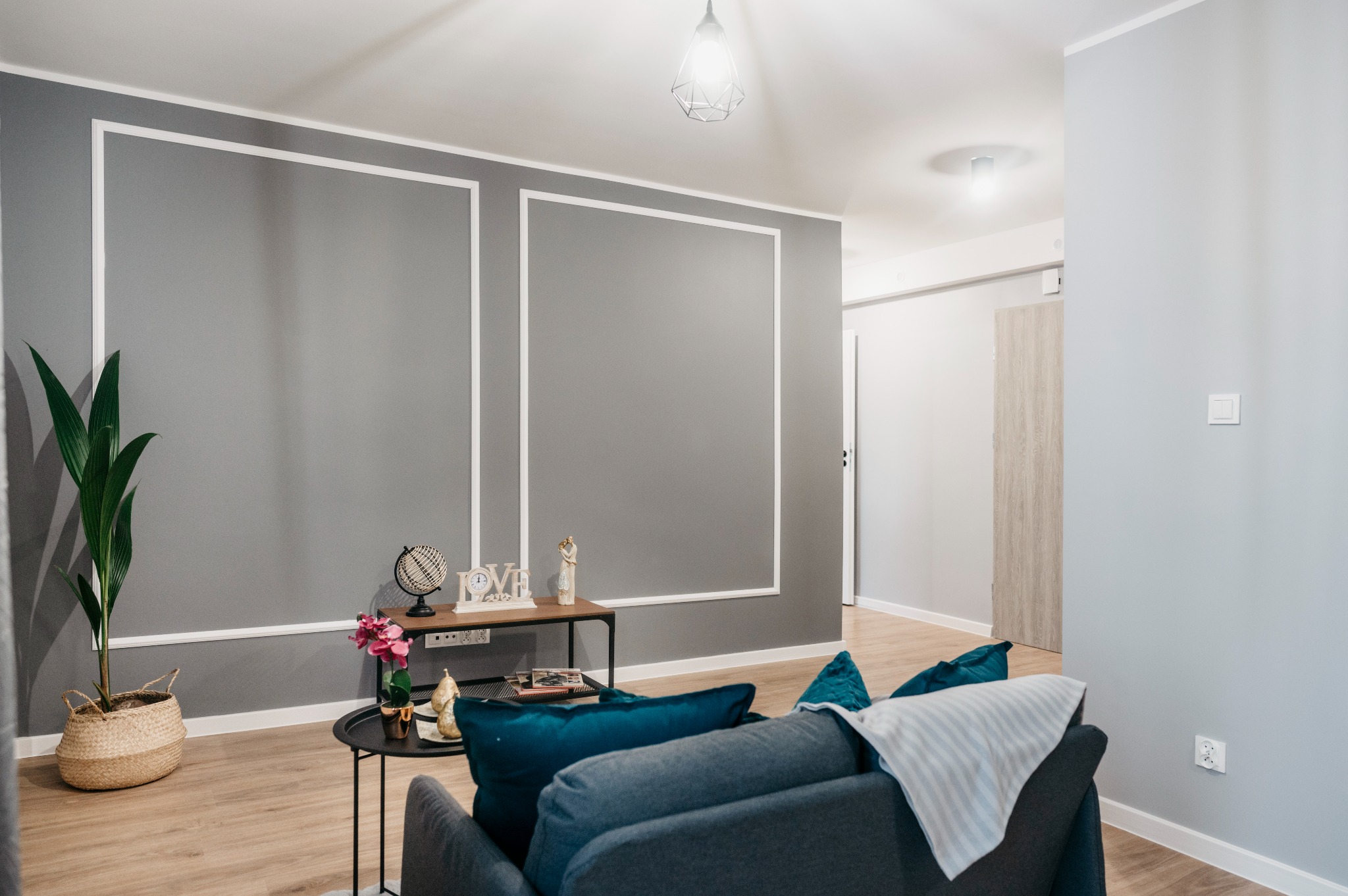Szare ściany z białymi dekoracyjnymi ramkami, drewniana podłoga, roślina w wiklinowym koszu, stolik z dekoracjami, fragment sofy z turkusowymi poduszkami i lampa sufitowa w nowoczesnym salonie.