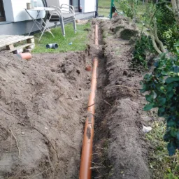 Pomarańczowa rura ułożona w wykopie ziemnym wzdłuż budynku, widoczne korzenie i fragmenty darni, w tle krzesła ogrodowe i butelka napoju.