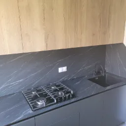 Nowoczesna kuchnia z drewnianymi szafkami górnymi i szarym blatem imitującym marmur, z czarną płytą gazową i zlewem w tym samym kolorze.