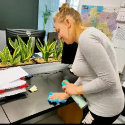 Kobieta w szarej bluzce spryskuje i przeciera niebieską ściereczką czarny blat biurka w biurze, na którym stoją doniczki z roślinami i monitory komputerowe, a w tle wisi mapa Polski.