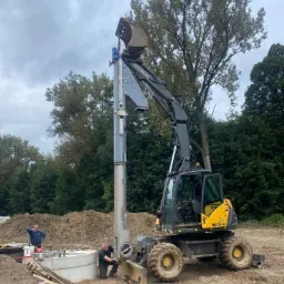 Żółto-czarna koparka Mecalac z zamontowanym pionowym urządzeniem do wbijania rur, dwóch mężczyzn przy betonowych kręgach, ziemia i drzewa w tle.