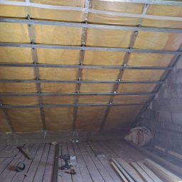 Krystian Drozdowski - Poddasze w trakcie ocieplania: widoczne metalowe profile konstrukcyjne, warstwa izolacji z folią, drewniana podłoga, narzędzia i materiały budowlane porozrzucane na podłodze.