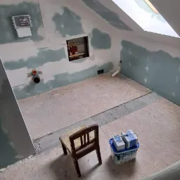 Poddasze w trakcie remontu: ściany z płyt kartonowo-gipsowych ze śladami szpachlowania, okno dachowe, odsłonięte rury instalacji, krzesło i wiadro z zaprawą.