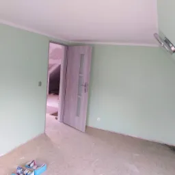 Świeżo pomalowane na jasnozielono ściany pokoju z otwartymi drzwiami, widoczne niedokończone podłogi i listwy przypodłogowe.