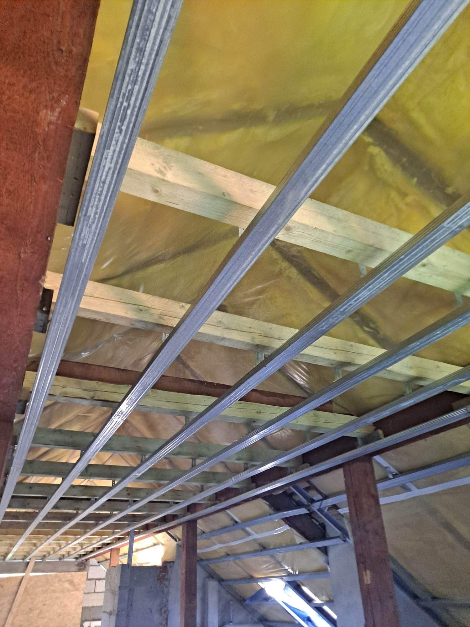 Metalowy szkielet sufitu podwieszanego zamontowany na drewnianej konstrukcji, widok z dołu w trakcie prac.